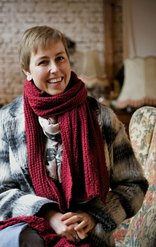 Anke lacht, je voelt haar ziel doorheen de foto. Ze zit in Arendonk op de bloemetjes sofa bij Tuintaferelen. Ze draagt een jas en sjaal.